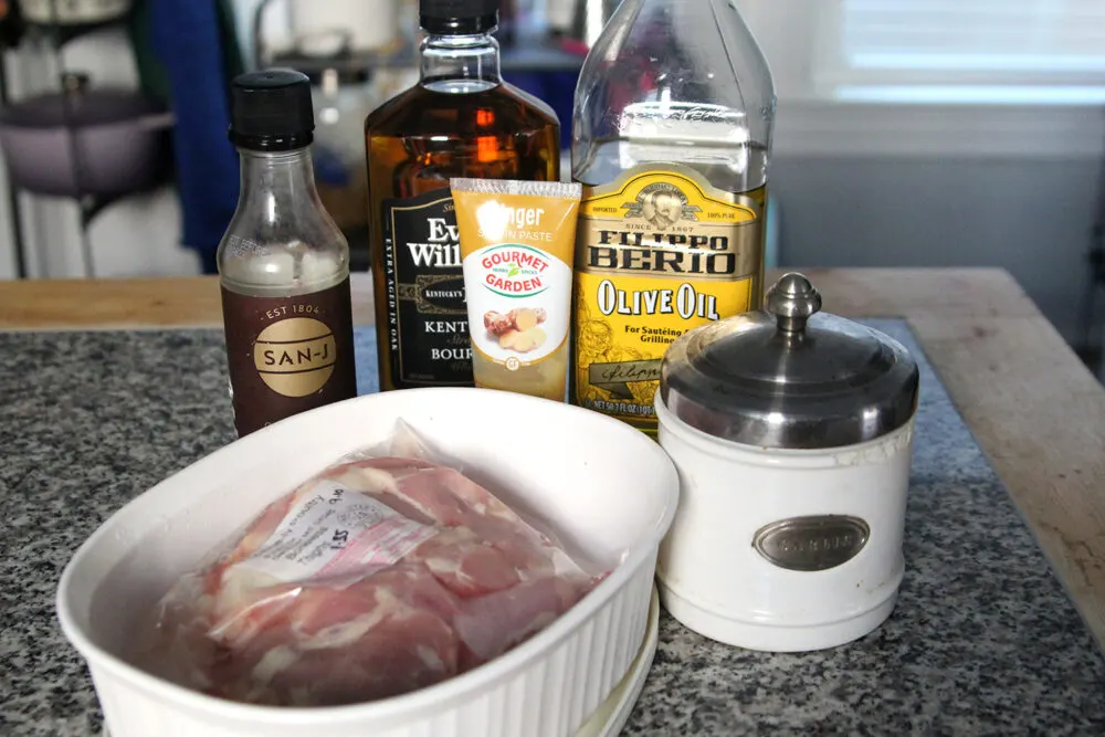 Soy sauce, bourbon, grated ginger, olive oil and chicken thighs are shown on a counter alongside a jar marked "garlic."