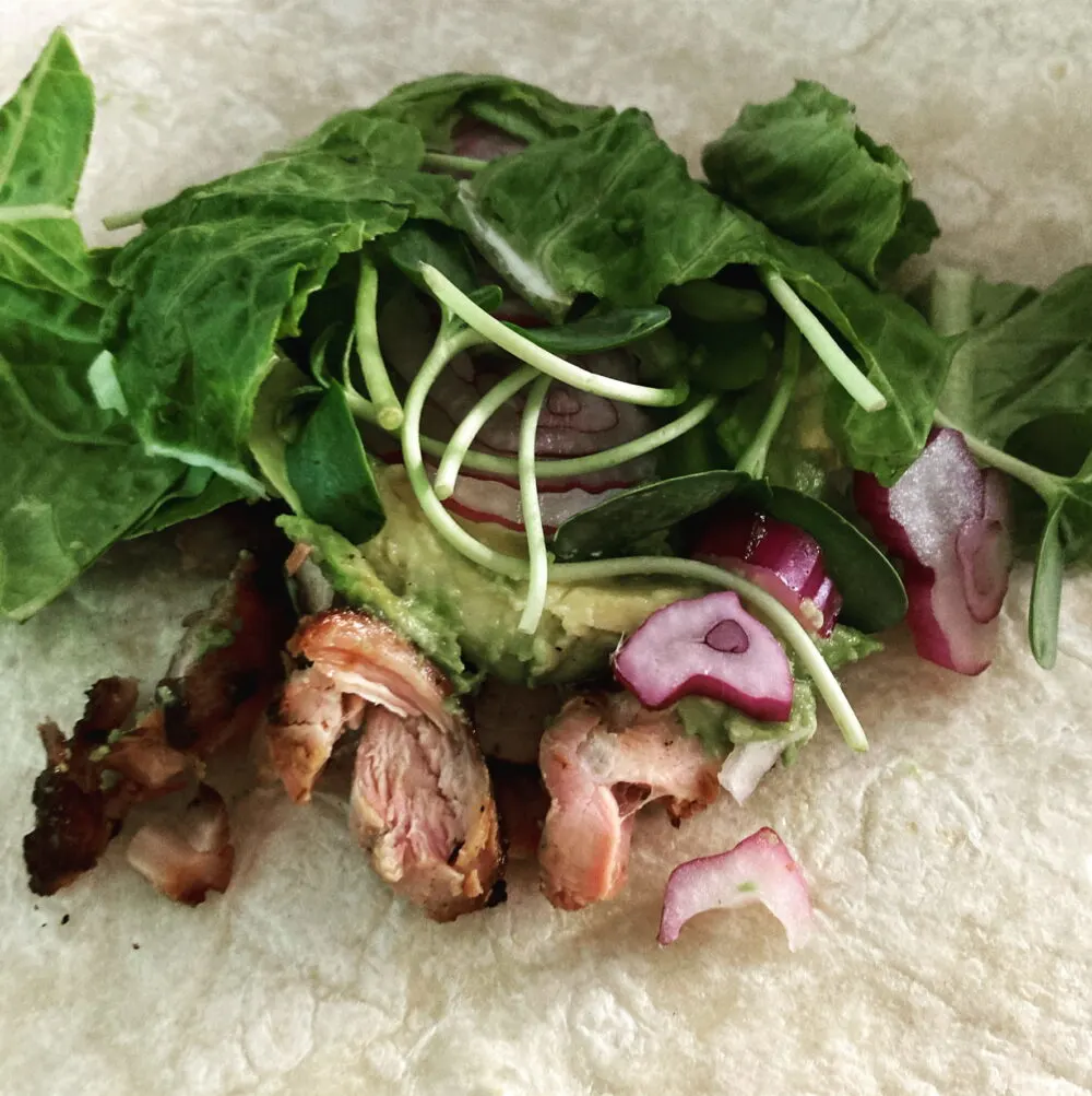 Chopped grilled chicken thighs are shown on a tortilla topped with avocado, sliced red onions and greens.