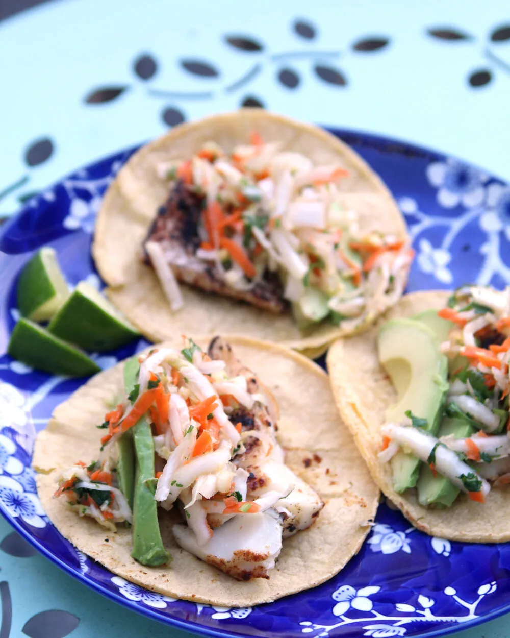Three fish tacos are laid out on a blue and white plate. The seasoned fish is topped with Cilantro Lime Slaw and avocado.