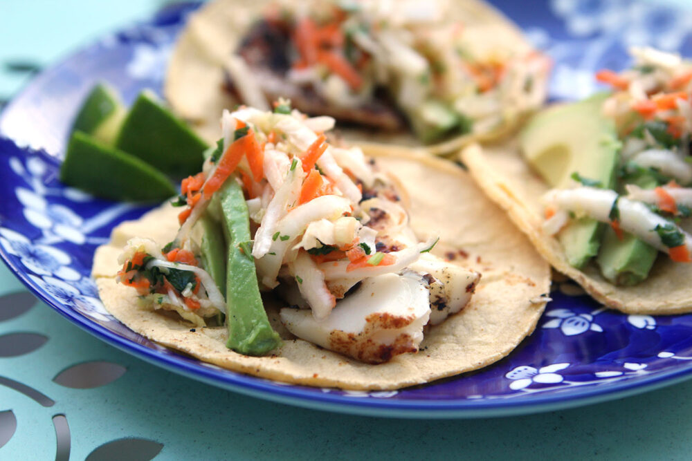 Three fish tacos are laid out on a blue and white plate. The seasoned fish is topped with Cilantro Lime Slaw and avocado.