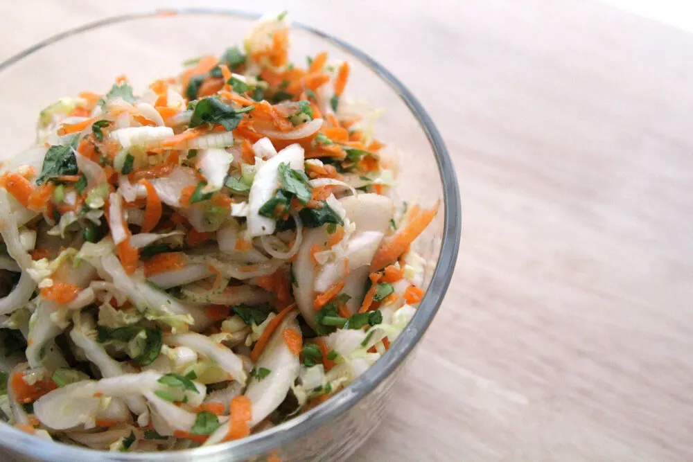 Cabbage, carrots, cilantro and onions are in a glass bowl on a counter, all mixed up.