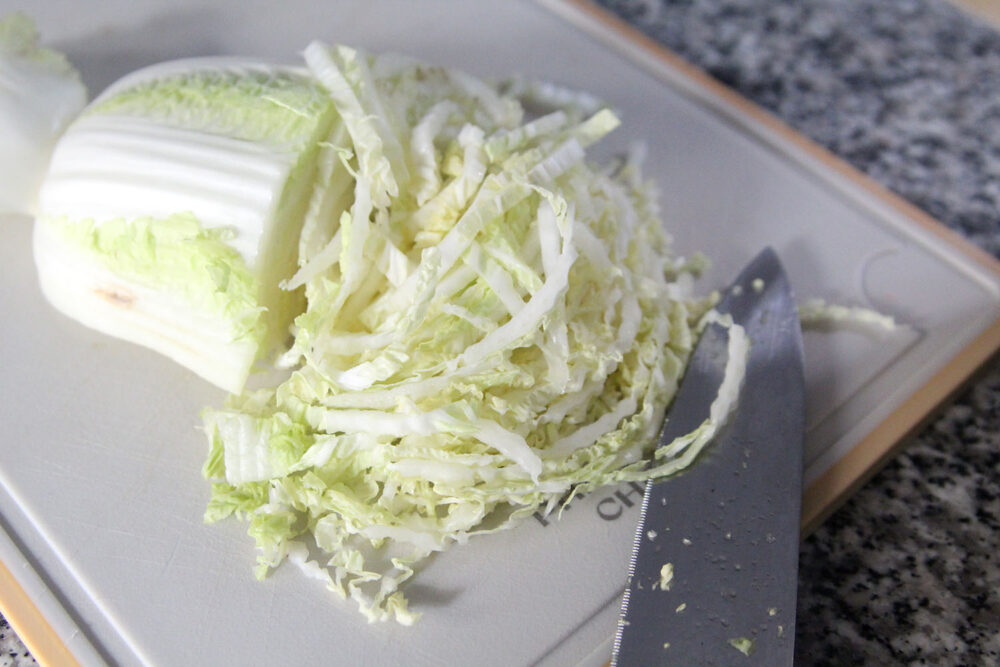 Napa cabbage is shown on a cutting board. Half of the head is finely shredded with a sharp knife laid nearby.