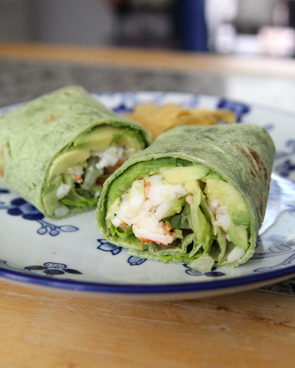 A wrap sandwich sits on a white and blue plate. Visible inside the wrap is green avocado, mostly white lobster, green lettuce and green basil.