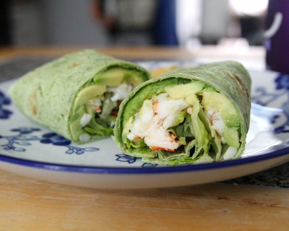 A wrap sandwich sits on a white and blue plate. Visible inside the wrap is green avocado, mostly white lobster, green lettuce and green basil.