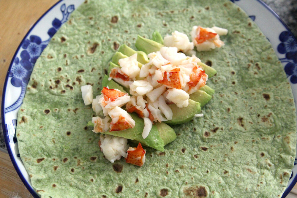 On a green wrap set on a white and blue plate, avocado slices are arranged with white and red lobster meat on top.