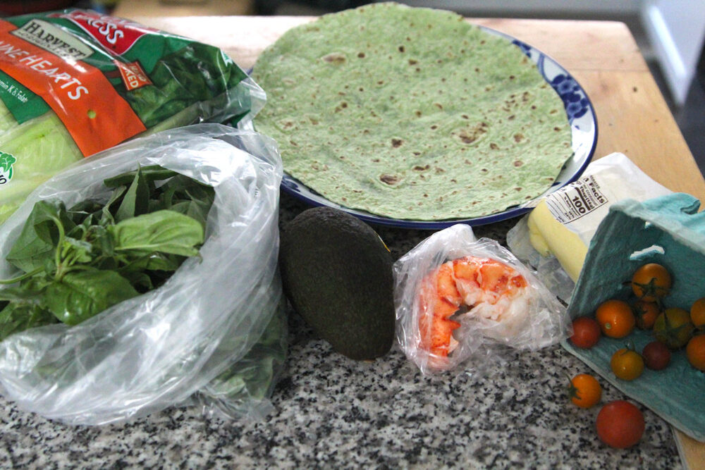 A collection of ingredients sit on a countertop including a bag of basil, a carton of cherry tomatoes, a whole avocado, a bag of lobster meat, a hunk of butter and a green wrap set on a plate. There's a bag of lettuce too.