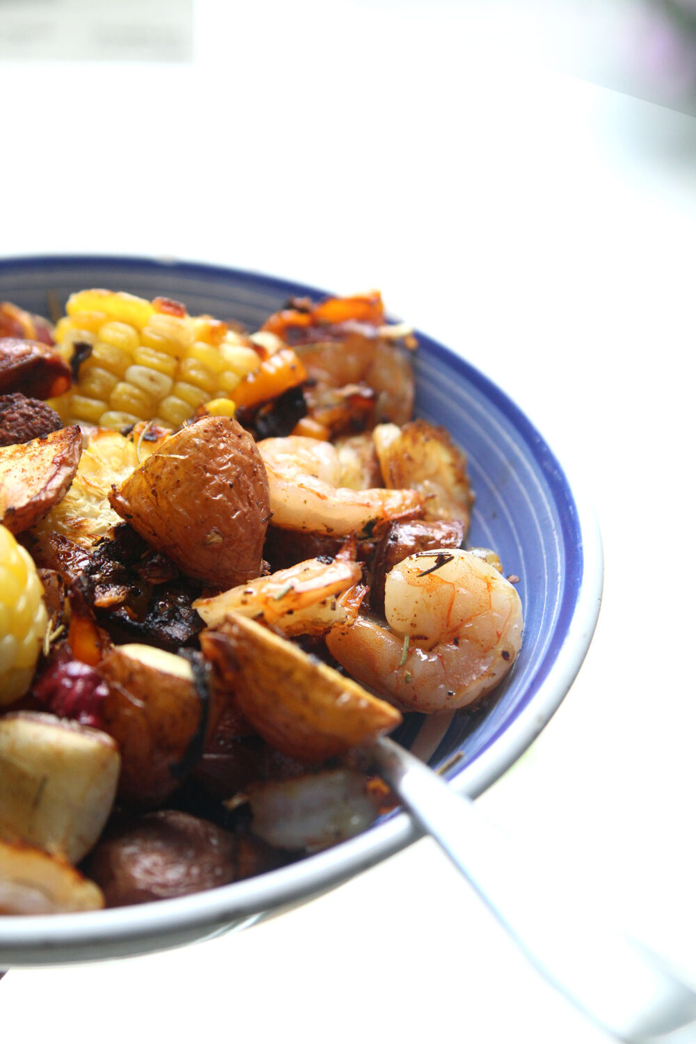 A blue and white plate holds a pile of potatoes, corn, shrimp, roasted onions and peppers. You can see green strips of rosemary on the dish as well.