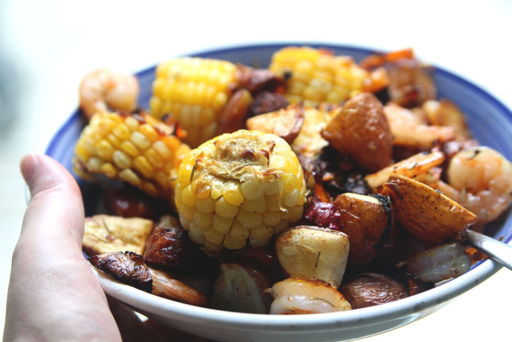 A blue and white plate is held in a visible hand. On the plate is a pile of potatoes, corn, shrimp, roasted onions and peppers. You can see green strips of rosemary on the dish as well.
