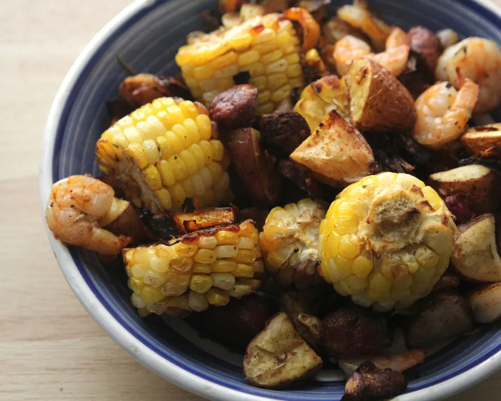 A blue and white plate is piled with potatoes, corn, shrimp, roasted onions and peppers. You can see green strips of rosemary and shrimps of red paprika on the dish as well.