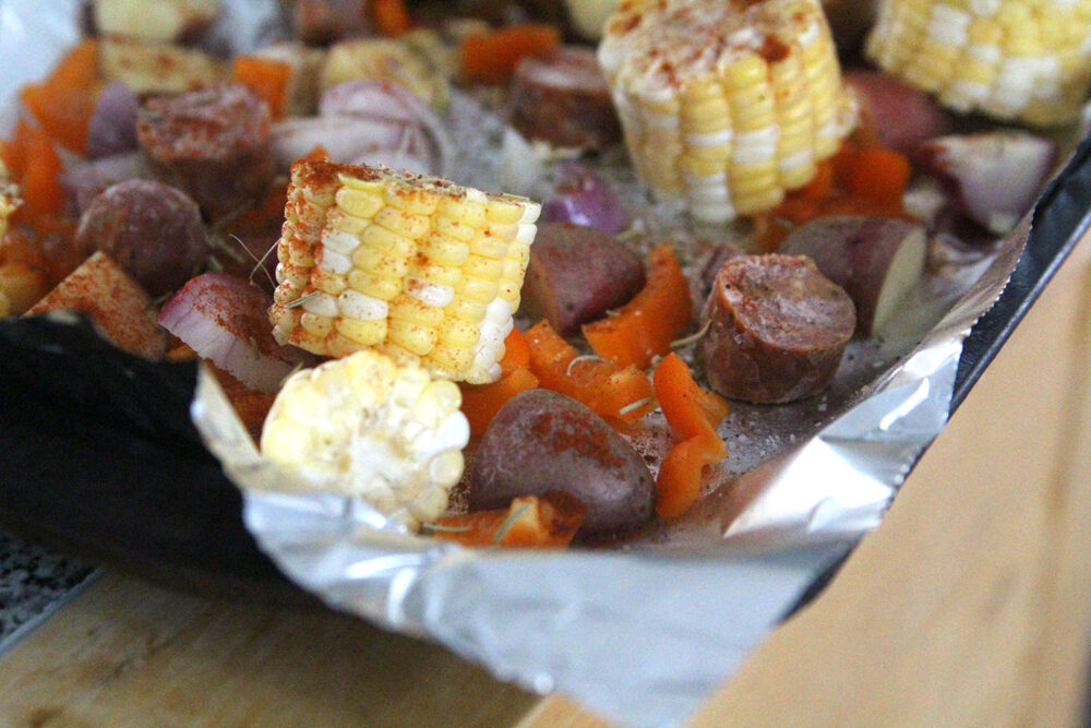 A baking sheet covered with foil holds corn, potatoes, red bell peppers, purple onions and sausage. Red paprika and light green rosemary are visibly sprinkled on the uncooked food.