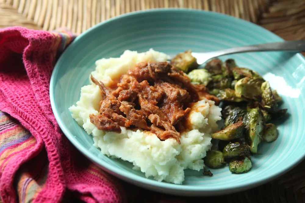 A blue plate is shown with mashed potatoes topped with reddish brown, saucy pulled pork. Brussels sprouts and a fork are also on the plate. It's sitting on a red cloth napkin on rattan.