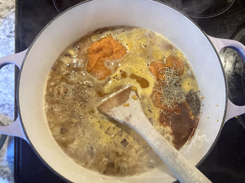 A pot of soup sit on a stove burner with visible herbs, pumpkin and onions in it.