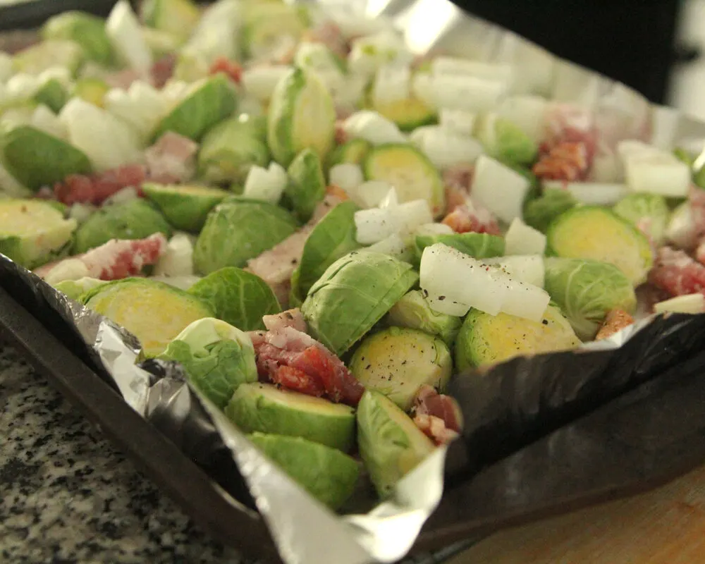 A baking sheet with Brussels sprouts, bacon and onions seasoned with salt and pepper sits on a counter.