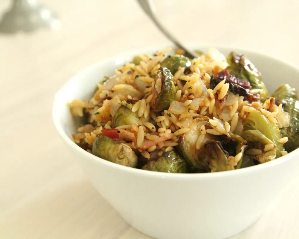 A white bowl filled with green and browned Brussels sprouts, dark brown bacon and yellow orzo sits on a white washed counter.
