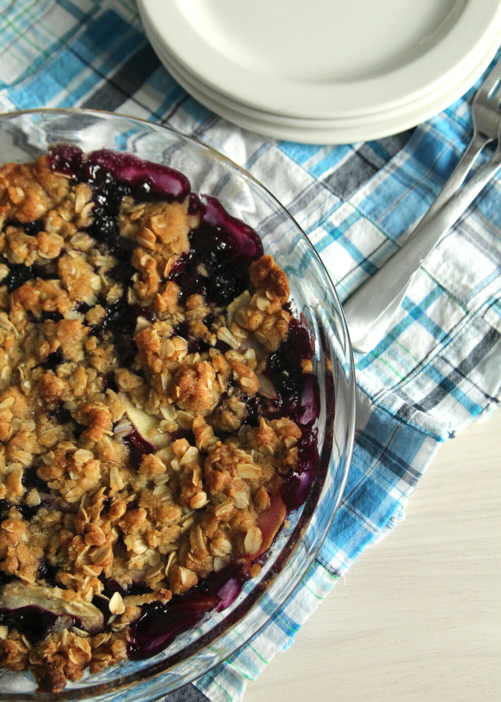 A purple mixture can be seen beneath a golden brown crumble in a clear pie plate set on a blue plaid fabric with white plates and forks nearby.
