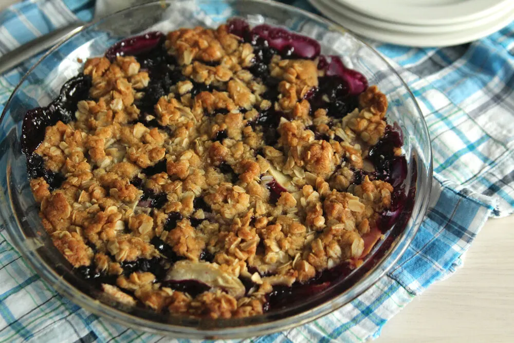 A purple mixture can be seen beneath a golden brown crumble in a clear pie plate set on a blue plaid fabric with white plates and a serving spoon nearby.