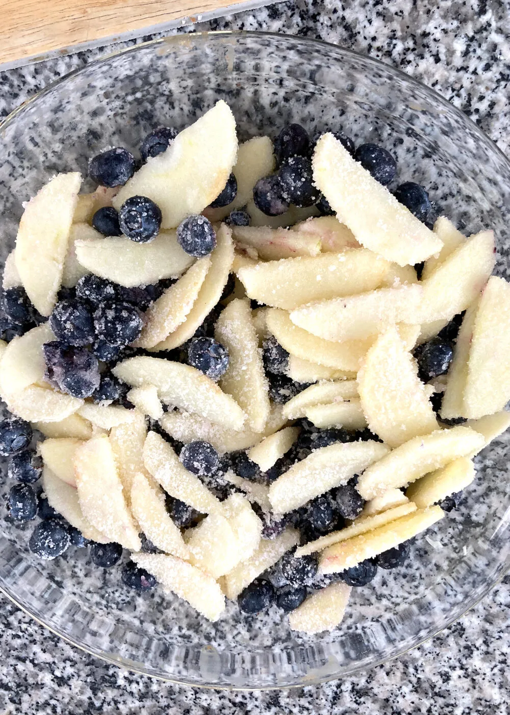 Blueberries and sliced apples dusted with sugar and flour can be seen in a clear bowl on a countertop.