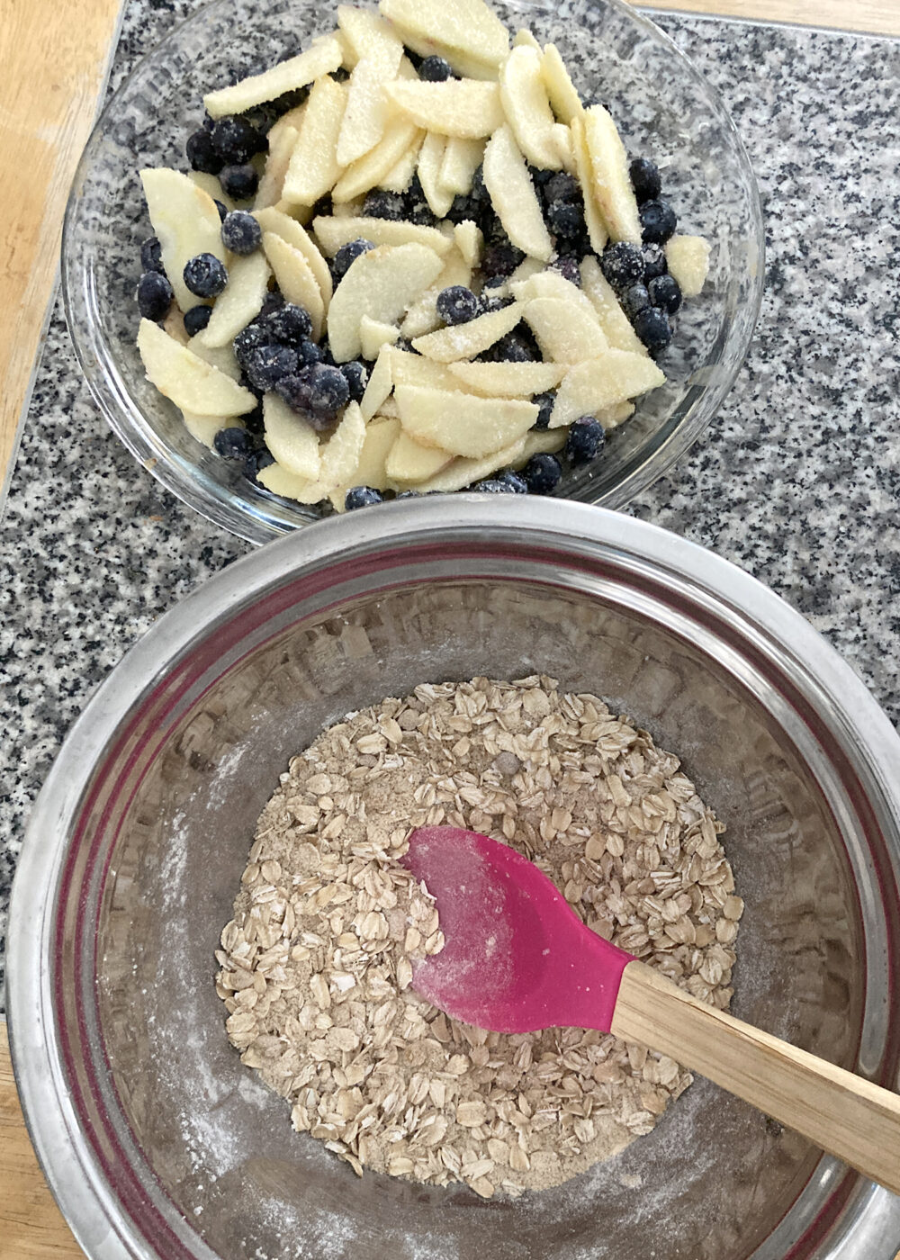 Two bowls sit side by side. In the upper portion of the frame, the glass bowl contains apple slices and blueberries mixed with sugar and flour. In the lower portion of the frame is a metal bowl with a oats mixture.