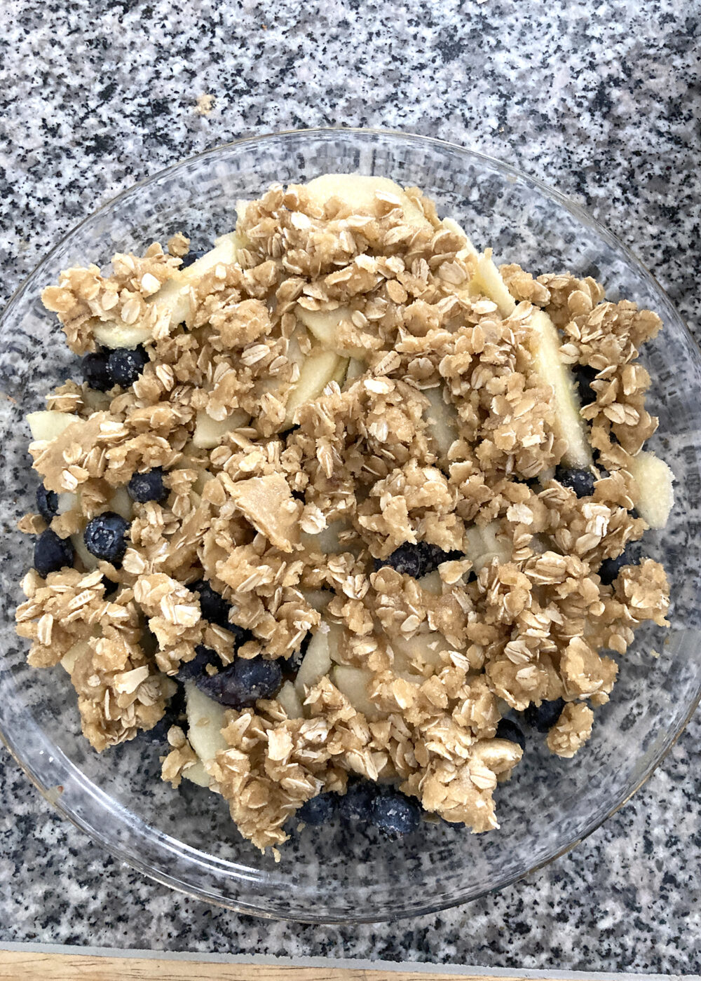 An unbaked Apple Blueberry Crisp sits in a clear glass pie plate on a countertop.