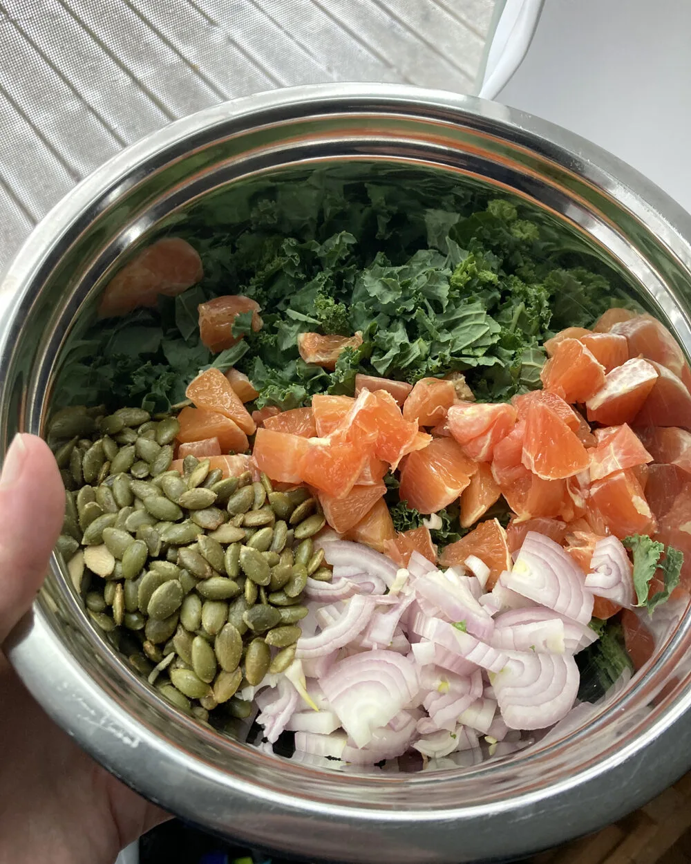 A metal mixing bowl with chopped shallots, pepitas, chopped kale and chopped cara cara oranges is shown.