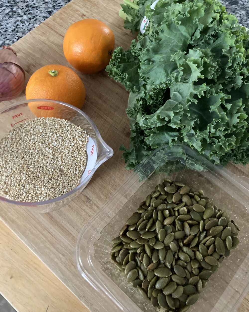Curly kale, two oranges, a shallot, a measuring cup with raw quinoa and a plastic container of pepitas are shown on a wooden cutting board.