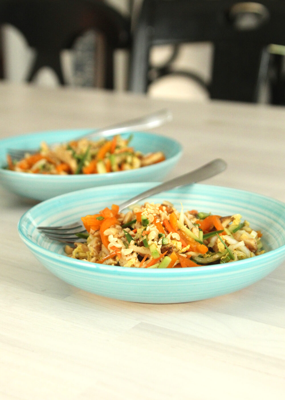 A salad made with lots of julienne cut veggies and a hearty addition of julienne cut chicken is shown in two teal bowls on a wooden surface. 