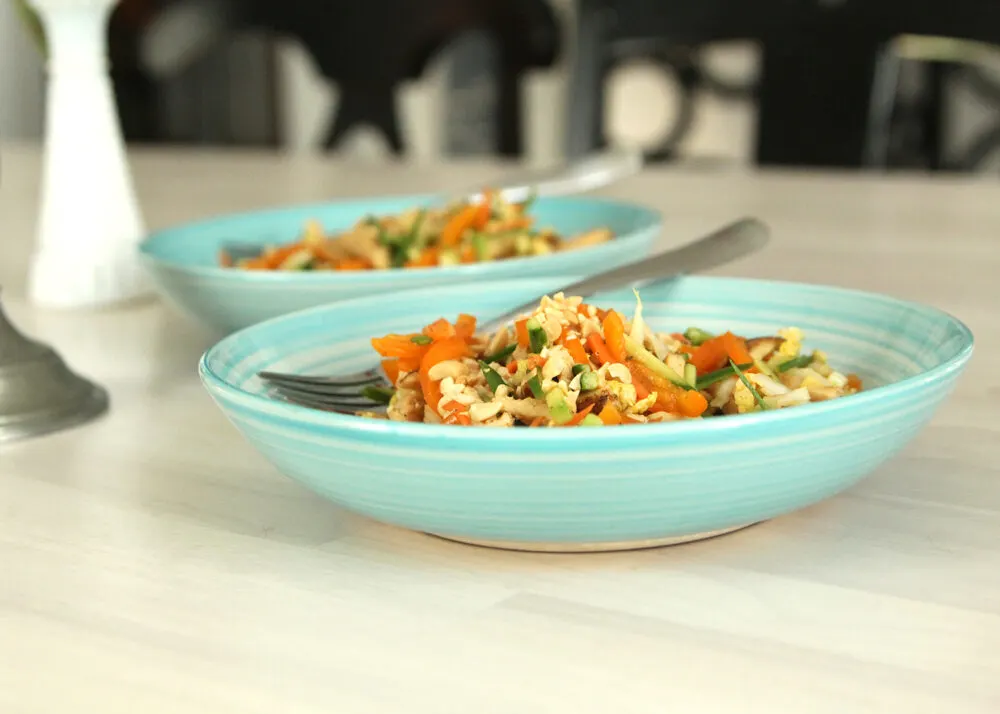 A salad made with lots of julienne cut veggies and a hearty addition of julienne cut chicken is shown in two teal bowls on a wooden surface. 