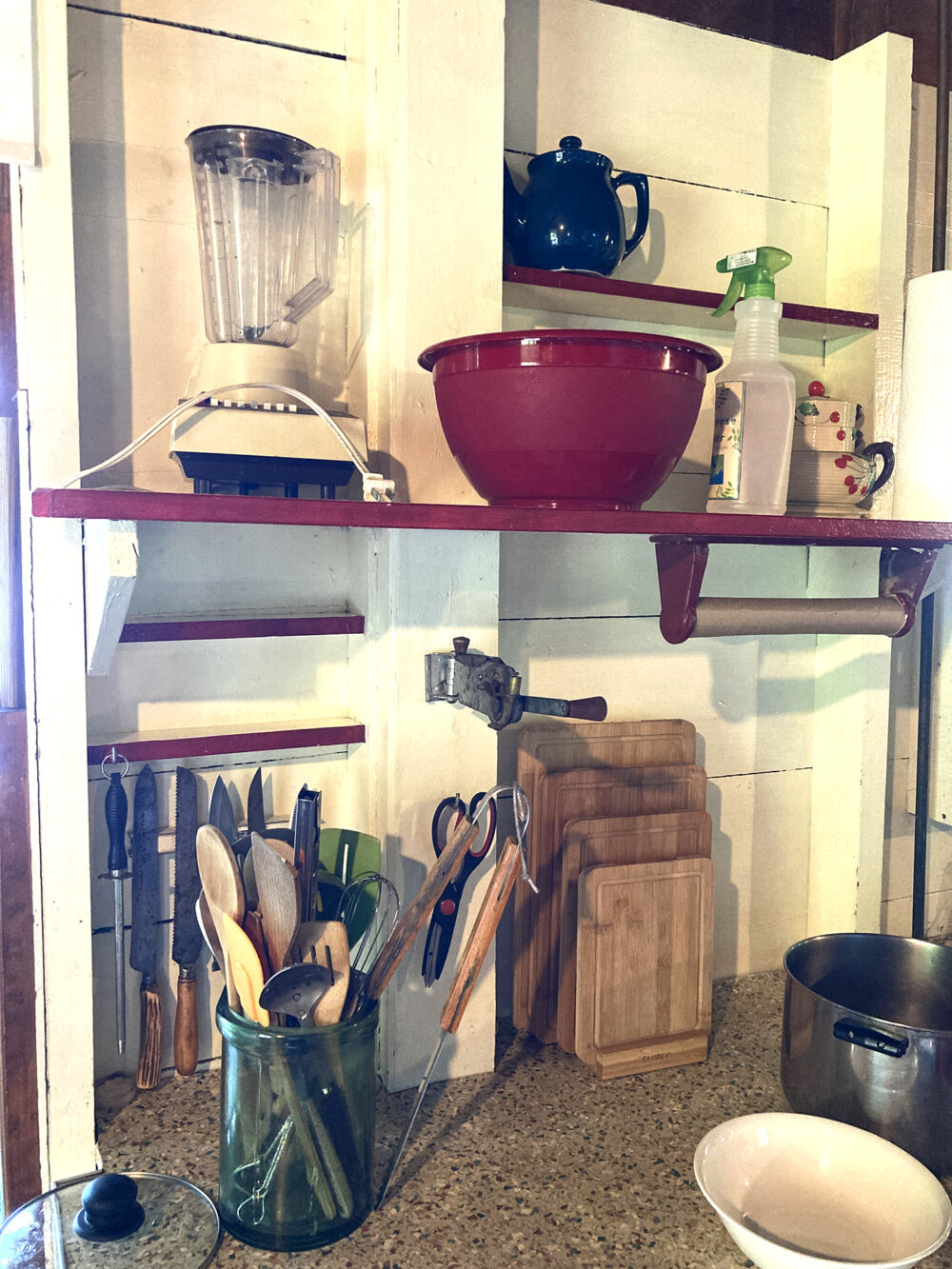 A countertop and white shelf is shown. Kitchen gear like a blender, utensils, knives and cutting boards are stored here.