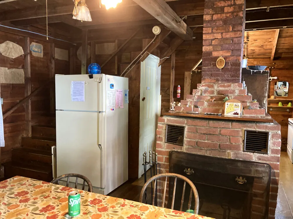 The inside of a rustic cabin is shown. A brick fireplace, a table and a refrigerator are shown.