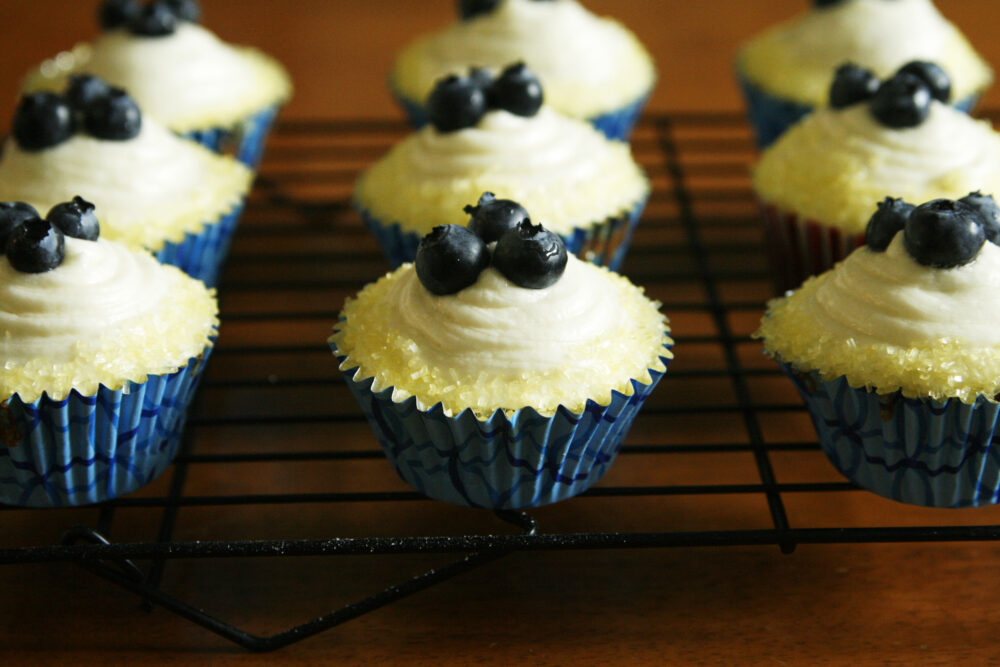 Cupcakes in blue wrappers sit on a cooling rack. They are frosted with buttercream, edged with yellow sugar crystals and each topped with three blueberries.