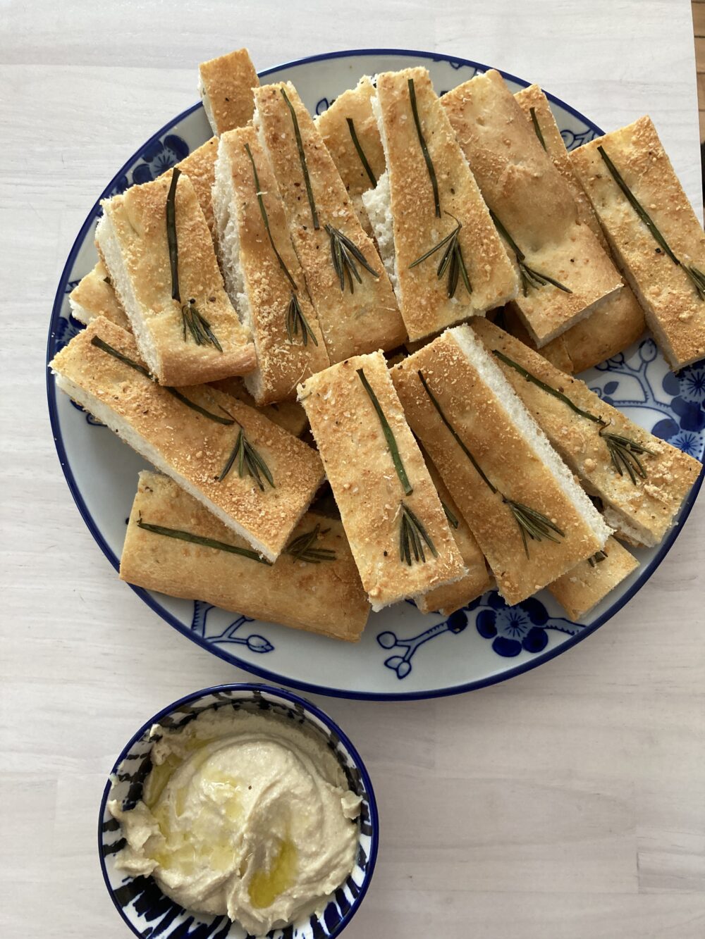 A plate of thin, long pieces of focaccia with a broom design comprised of rosemary and chives is shown on a white and blue plate with a bowl of hummus nearby.