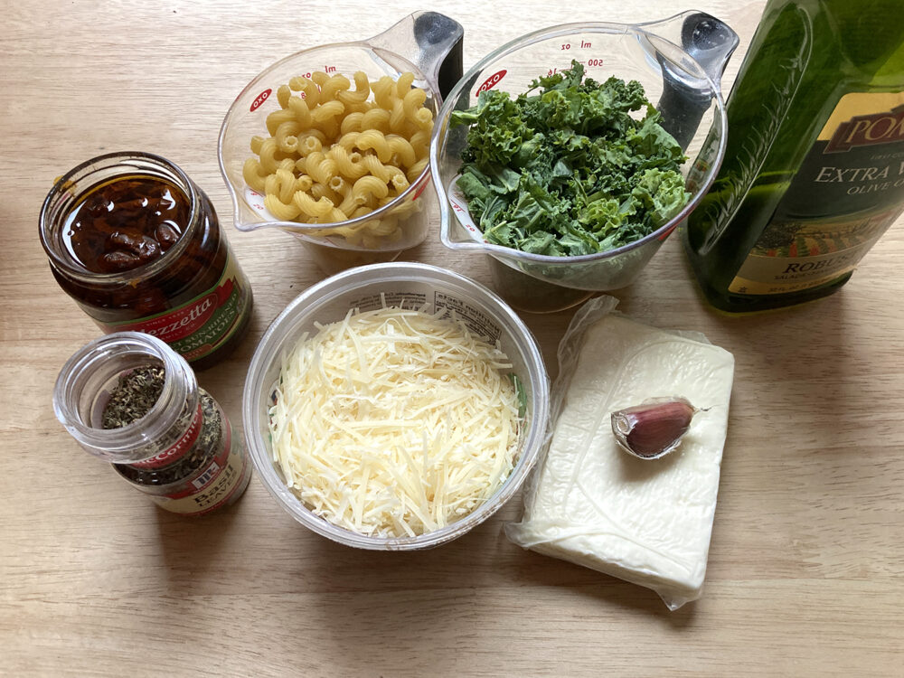 A jar of sundried tomatoes, a container of dried basil, a measuring cup with dried pasta, a container of shredded parmesan, a measuring cup of chopped kale, a block of feta, a clove of garlic and a bottle of olive oil is shown on a countertop.