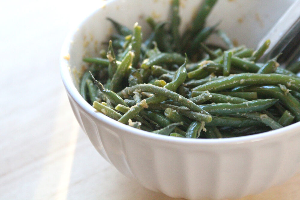 A white bowl containing green beans flecked with bits of roasted garlic sits on a wooden countertop.