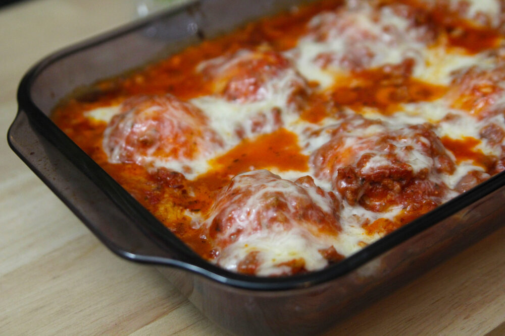 A purple glass baking dish filled with red sauce, white cheese and lumps of stuffed shells is shown on a wooden surface.