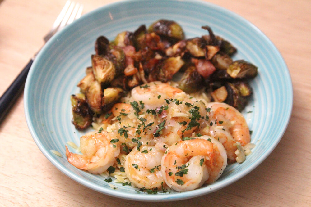A blue shallow bowl holds a pile of pink shrimp with bits of shallots and garlic and a sprinkle of parsley. Roasted Brussels sprouts can be seen in the background.