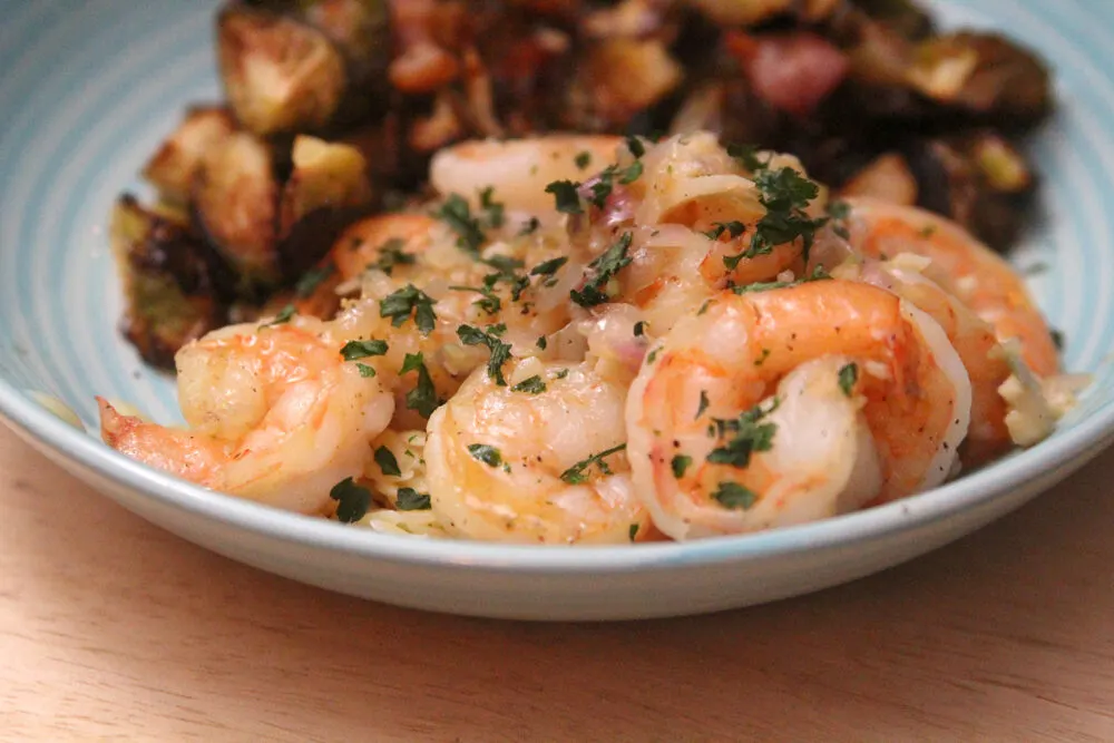 A blue shallow bowl holds a pile of pink shrimp with bits of shallots and garlic and a sprinkle of parsley. Roasted Brussels sprouts can be seen in the background.