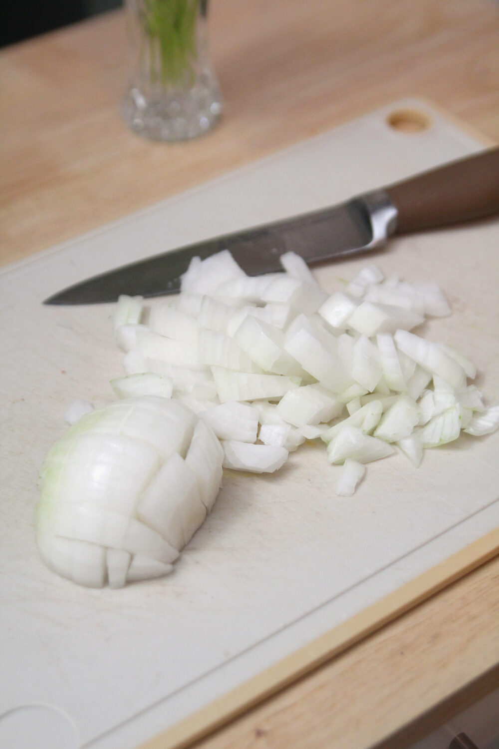 A chopped onion is shown on a cutting board next to a knife. Half of the onion is cut but in a half-moon shape and the rest is in a pile.