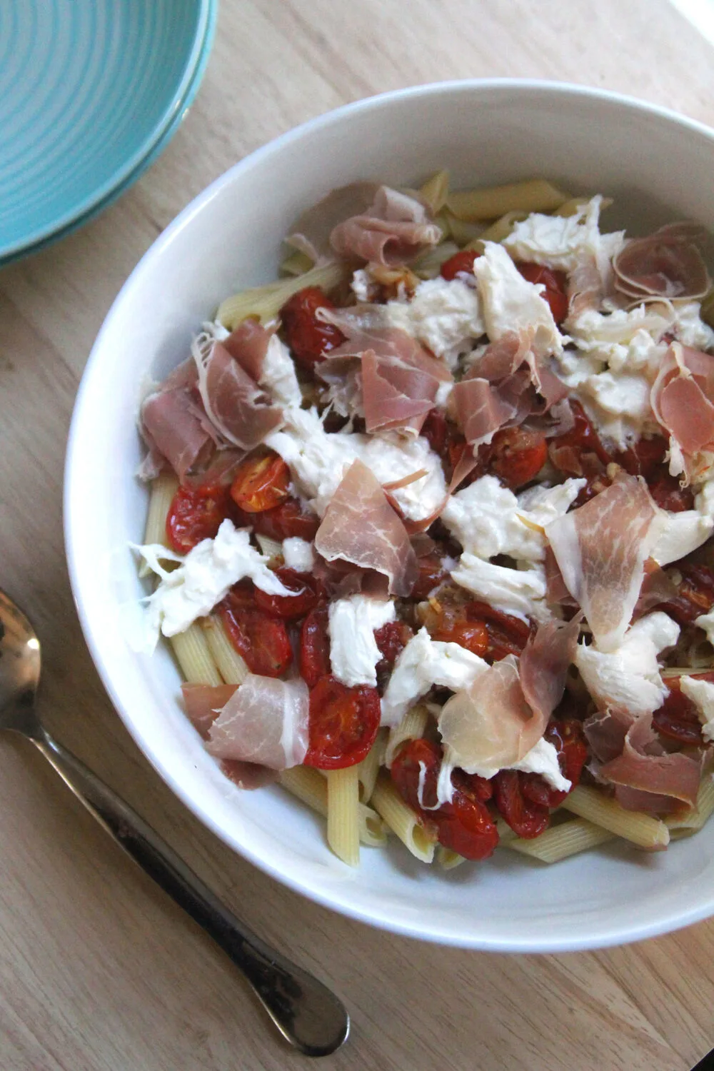 A white bowl is on a wooden surface with turquoise bowls nearby. The bowl contains red roasted tomatoes, white burrata cheese, pink prosciutto and yellow penne pasta.