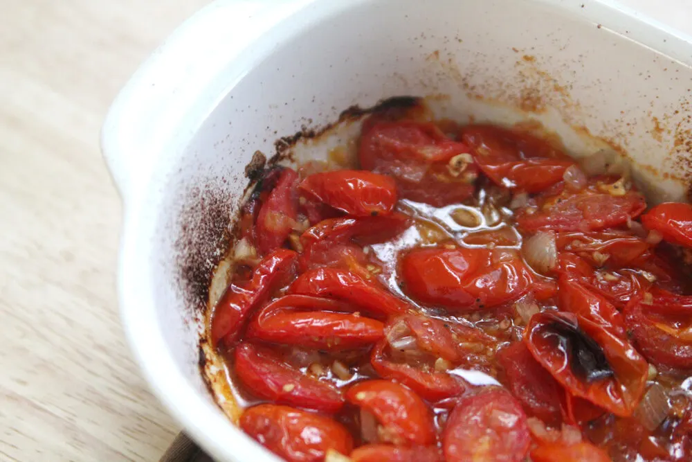 A white baking dish is seen after baking with bright red tomatoes sitting in juices. There are dark parts around the edges of the dish.