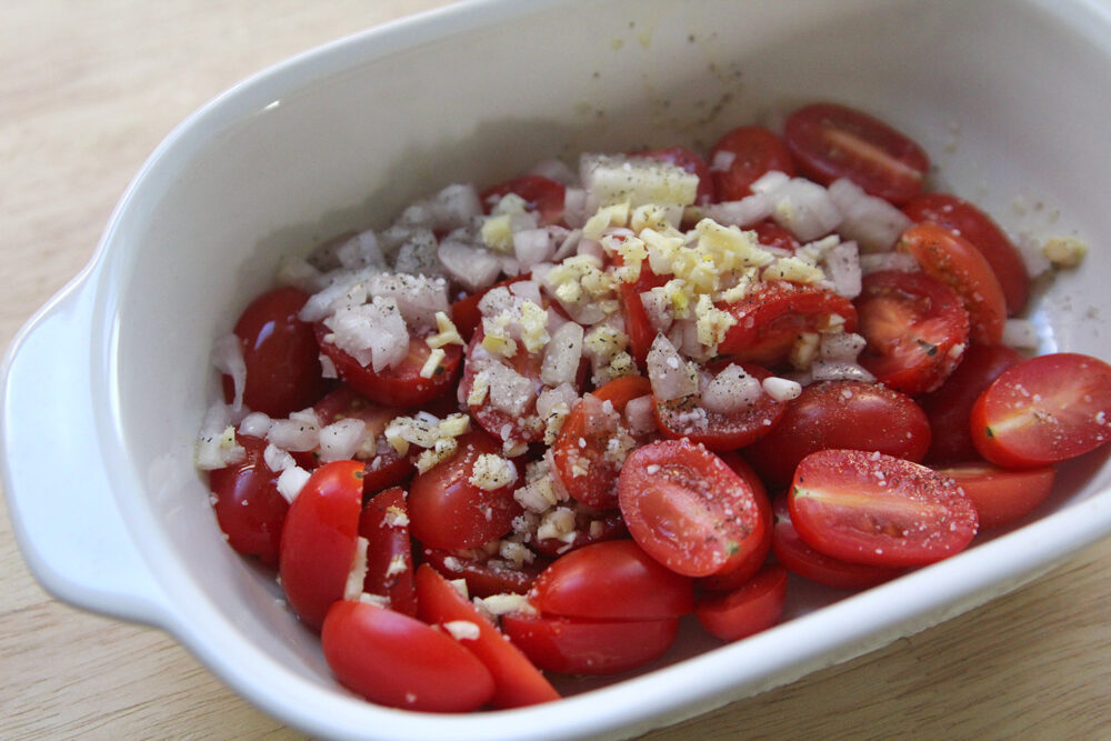 Red tomatoes are seen in a white oblong baking dish topped with tiny bits of shallots and garlic with visible salt and pepper.