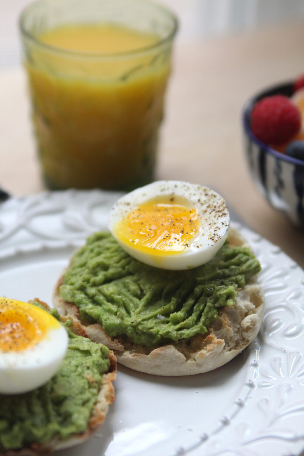 A white plate is shown with two English muffin halves topped with mushed avocado and soft boiled eggs that have been seasoned with salt and pepper. A glass of orange juice is shown in the background.