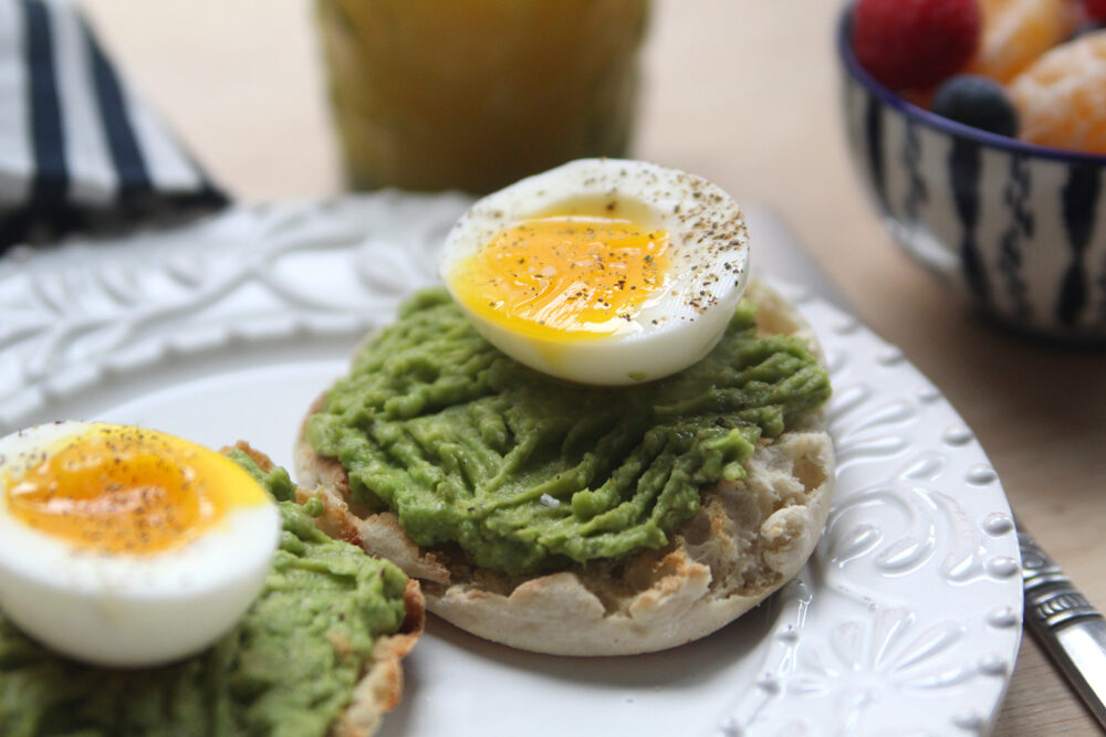 A white plate is shown with two English muffin halves topped with mushed avocado and soft boiled eggs that have been seasoned with salt and pepper.