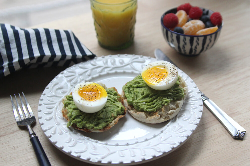 A white plate is shown with two English muffin halves topped with mushed avocado and soft boiled eggs that have been seasoned with salt and pepper. A fork, knife, stripped napkin, glass of orange juice and bowl of fruit are shown nearby.