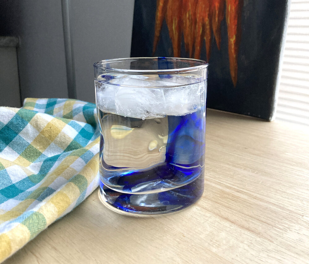 A glass of ice water is shown on a counter with a kitchen towel nearby and a painting of carrots in the background.