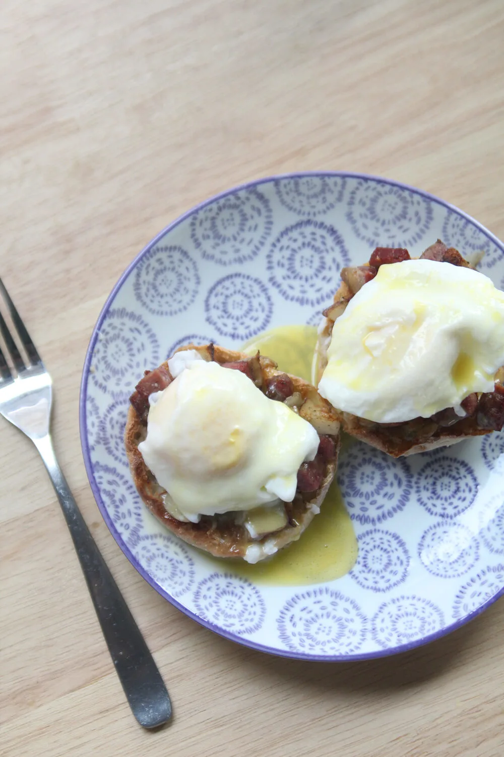 A plate with a purple and white design is shown with two light brown English muffin halves topped with brown corned beef hash, white eggs and yellow hollandaise sauce. A fork sits nearby.