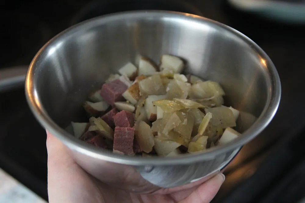 A silver bowl is held in a hand. It contains reddish-brown cubes of meat, yellowish pieces of onion and whiteish pieces of potato.