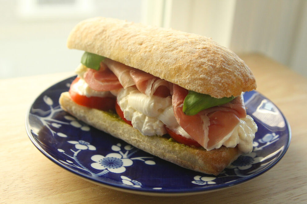 A sandwich on tan ciabatta bread is shown on a blue and white plate sitting on a wooden countertop. It has red tomatoes, white burrata cheese, pink prosciutto and green basil visible.