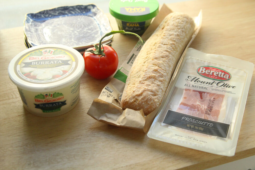 Ingredients for a Burrata Caprese Sandwich are shown on a wooden countertop.