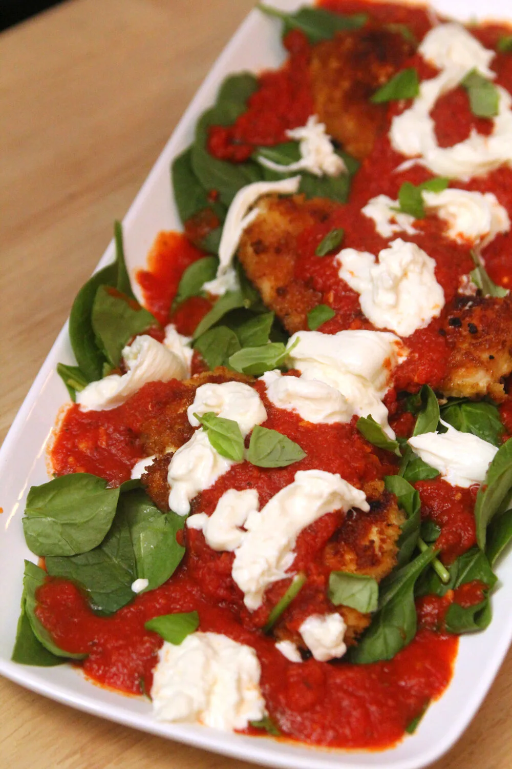 A white platter on a light brown wooden table holds green spinach topped with red marinara sauce, brown breaded chicken cutlets, white burrata cheese and green basil.