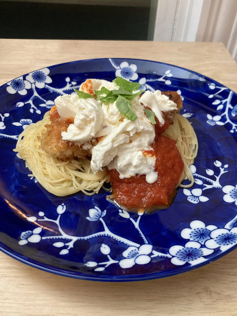 A blue and white flowered plate on a light brown wooden surface holds thin spaghetti, brown breaded chicken cutlets, red marinara sauce, white burrata cheese and green basil.